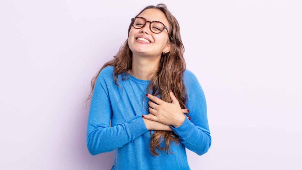 a girl in blue wearing spectacles smiling and having both her hands on her heart.